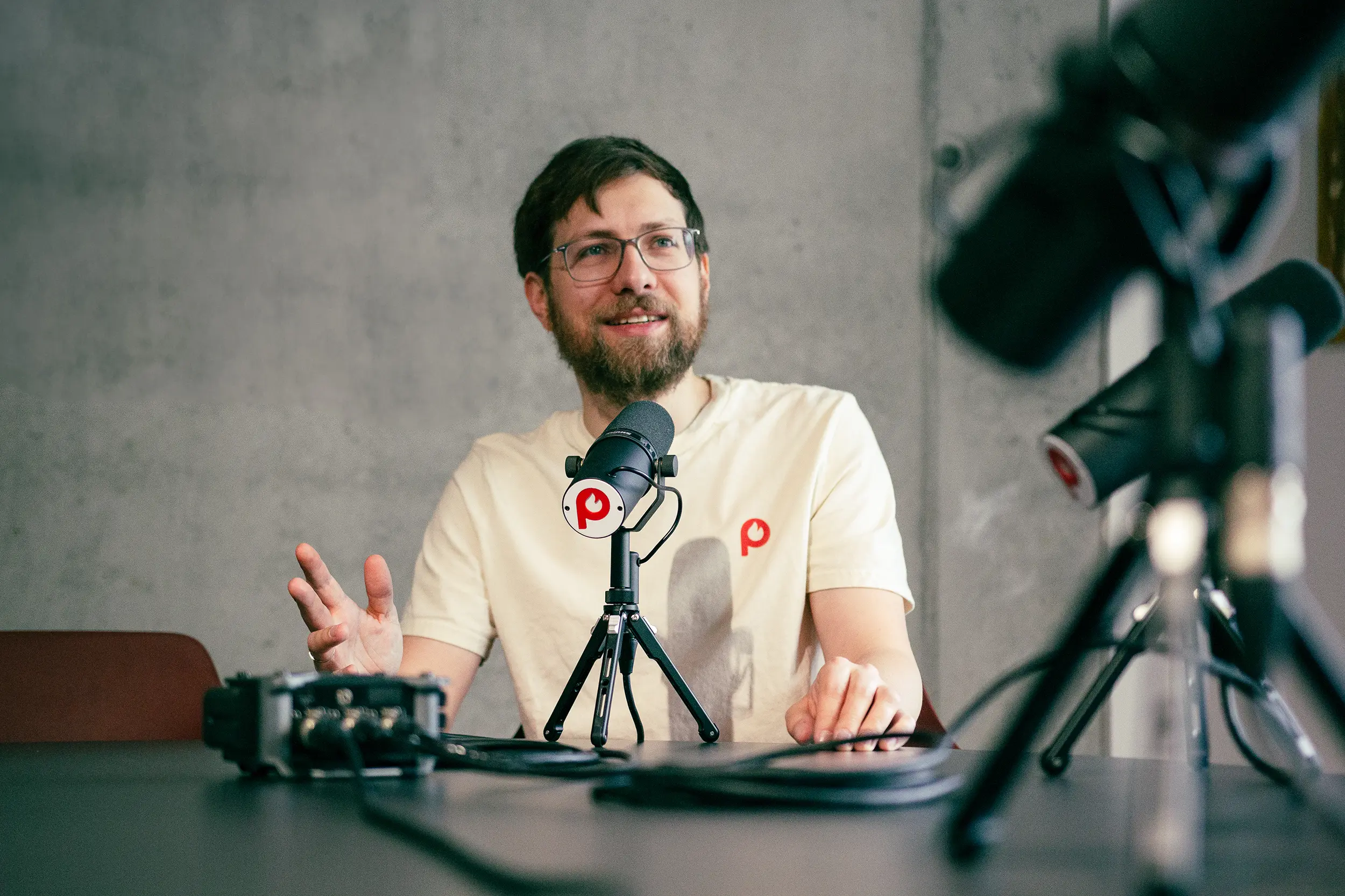 pepper-pods-header-chris-schmid Chris Schmid, Gründer von Pepper Pods bei der Aufnahme mit einem schwarzen Tischmikrofon sitzend an einem Tisch mit mehreren unscharfen Mikrofonen und weiterem Podcast Equipment im Vordergrund